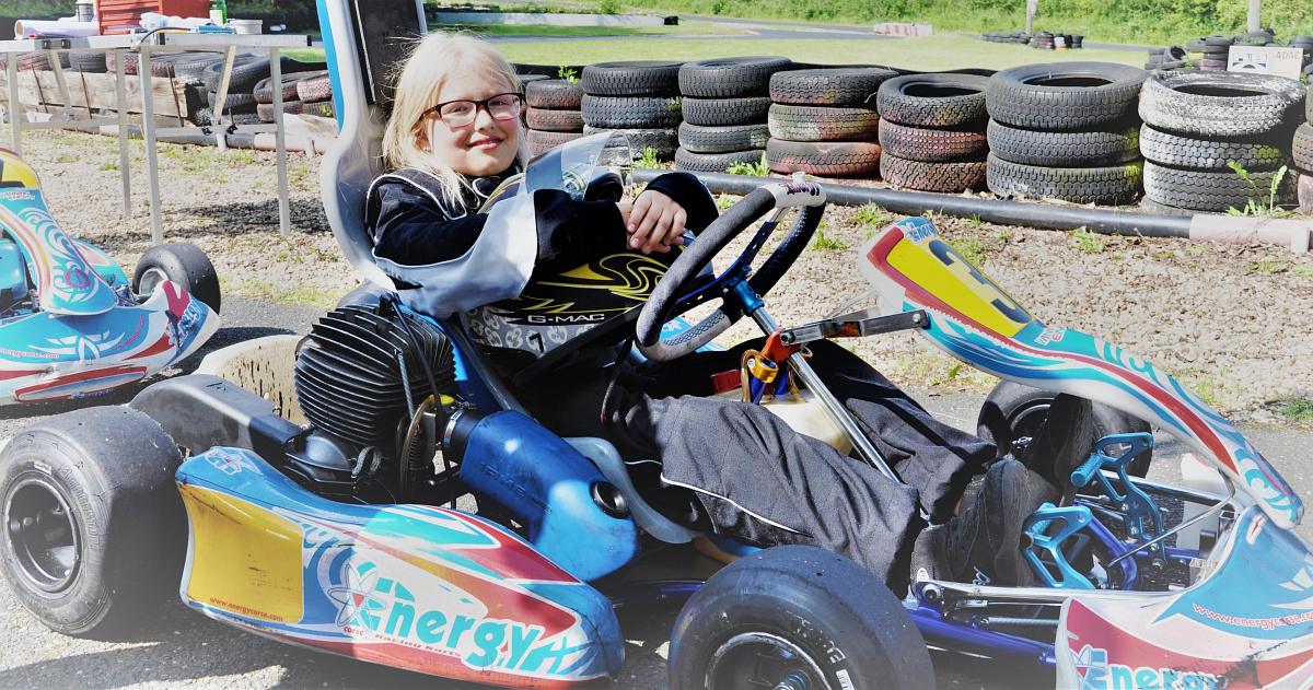 Blondes Mädchen sitzt in Gokart in voller Rennmontur und hält ihren Helm fest.