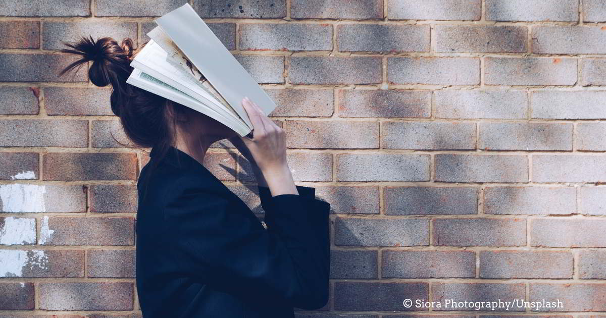 Junge Frau mit Hochsteckfrisur hält sich ein aufgeschlagenes Buch vors Gesicht