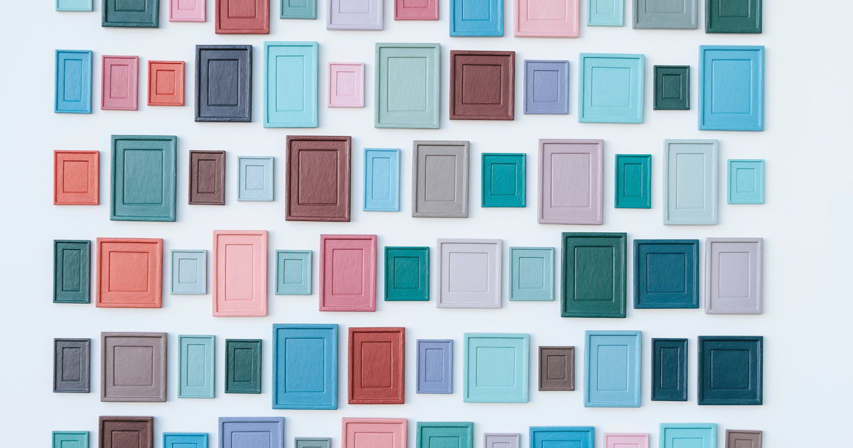 In pink, türkis, helblblau, braun und grün angemalte Bilder verschiedener Größen dicht an dicht an eine weiße Wand gehängt