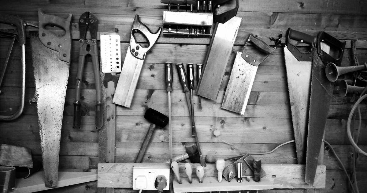 Schwarz-weiß-Foto von Werkzeugen wie Sägen, Hammer, Schraubenzieher und ähnliches, die an einer Holzwand hängen.