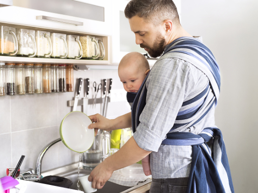 Andere Bildsprache: Ein Mann mit einem Baby im Tragetuch spült Geschirr in der Küche