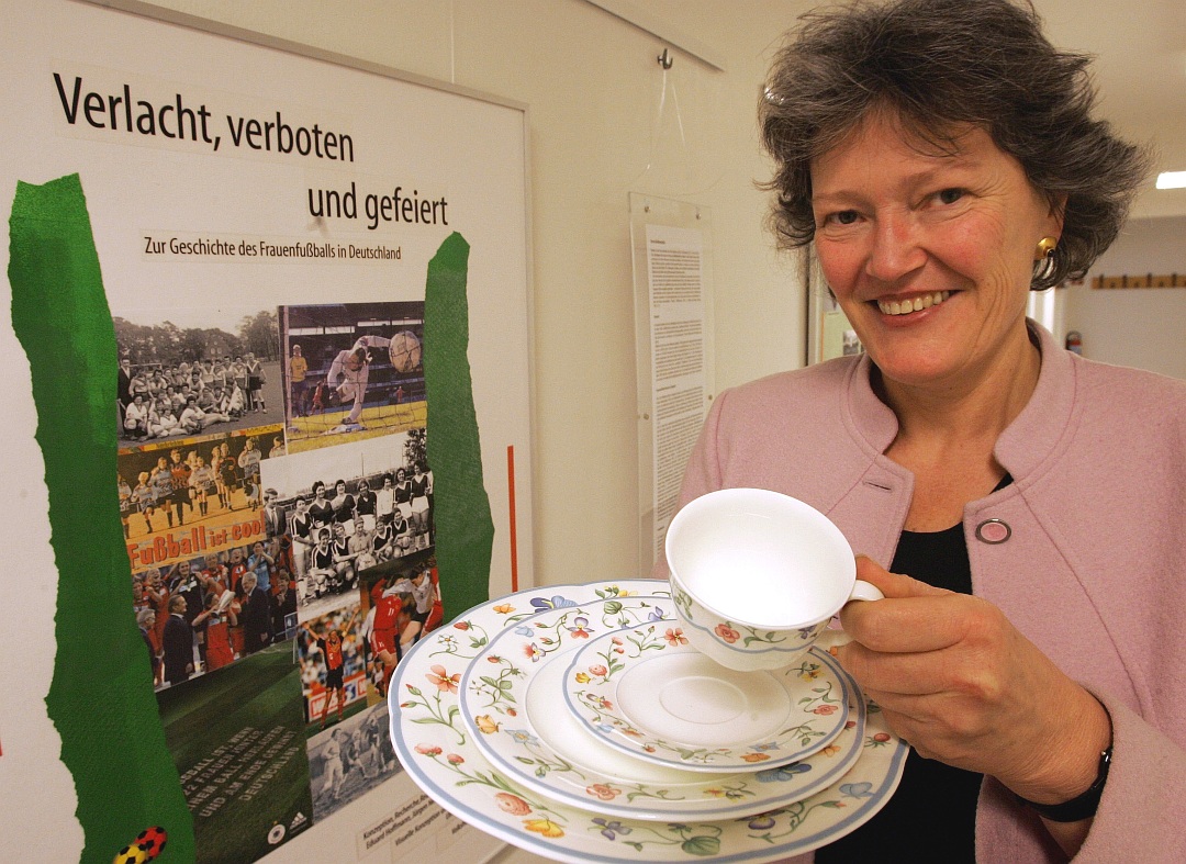 Frau steht vor einem Plakat zur Geschichte des Frauenfußballs und zeigt ein buntgeblümtes