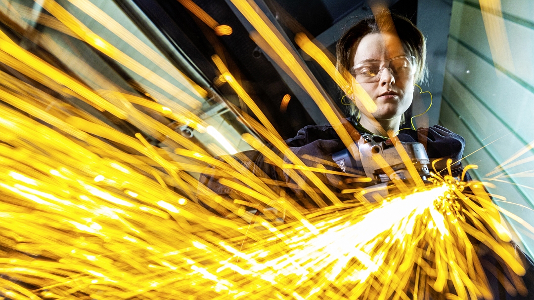 Frau mit Schutzbrille arbeitet mit einer Flex. Die Funken sprühen goldgelb.