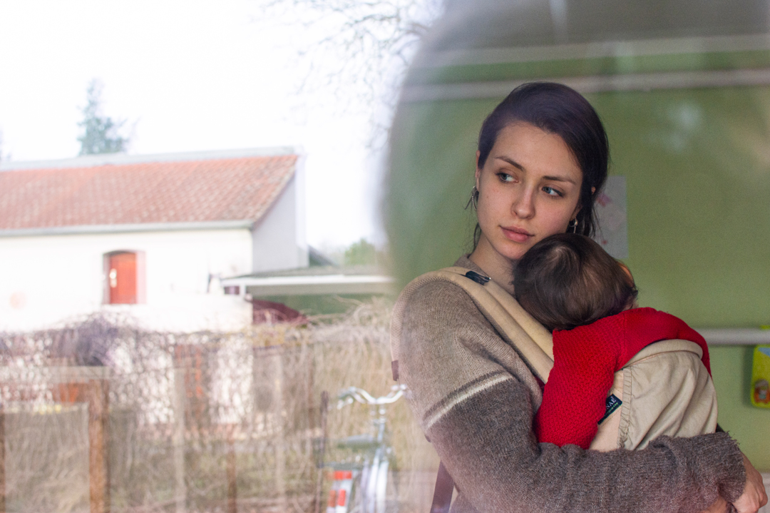 Blick von außen durch ein Fenster eines Frauenhauses. Drinnen steht eine junge Frau, sie hält ein Baby. Sie sieht nachdenklich aus. In der Fensterscheibe spiegelt sich ein weiteres Haus.