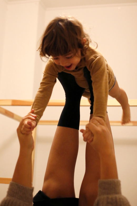 Ein etwa dreijähriges Kind mit braunen Locken schwebt über ihrer Mutter in der "Fliegerposition": sie ballanciert auf dem Rücken liegend das Kind auf ihren Füßen.