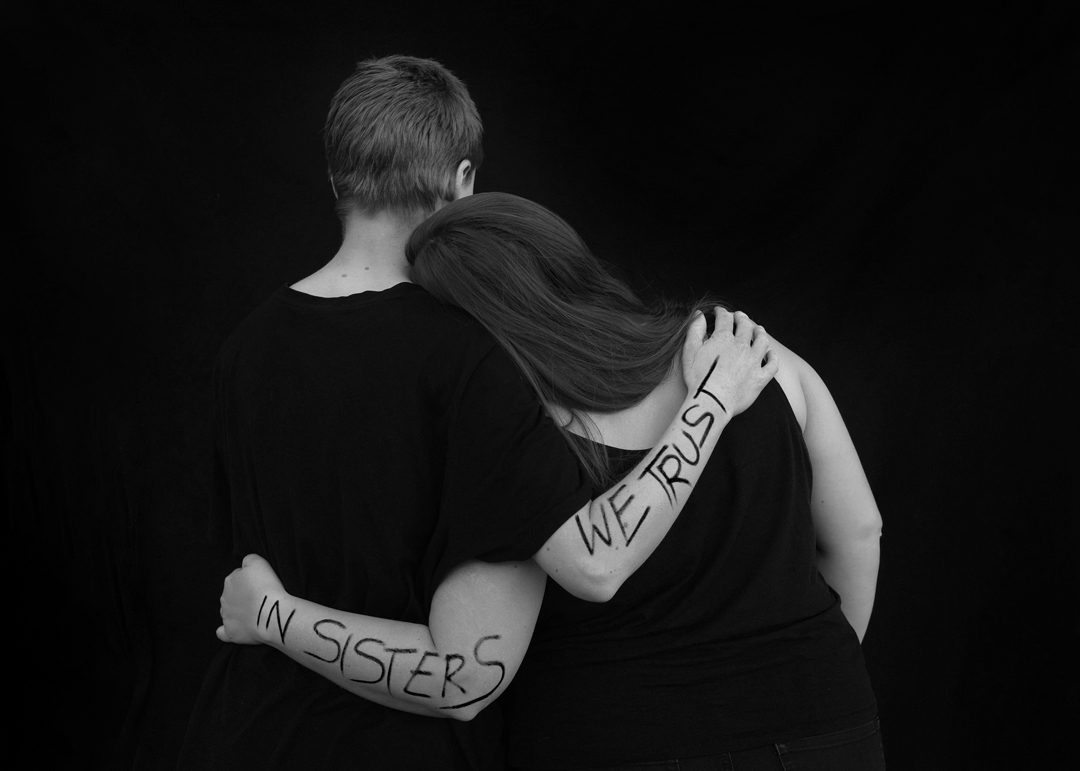 Schwarz-weiß-Foto. Rückenansicht: zwei Frauen in schwarzer Kleidung haben die Arme um Schulter und Hüfte gelegt. Auf den Unterarmen steht mit schwarzer Schrift: in sisters we trust