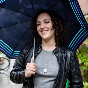 Frau mit dunklen Locken trägt einen aufgespannten Regenschirm und lacht in die Kamera