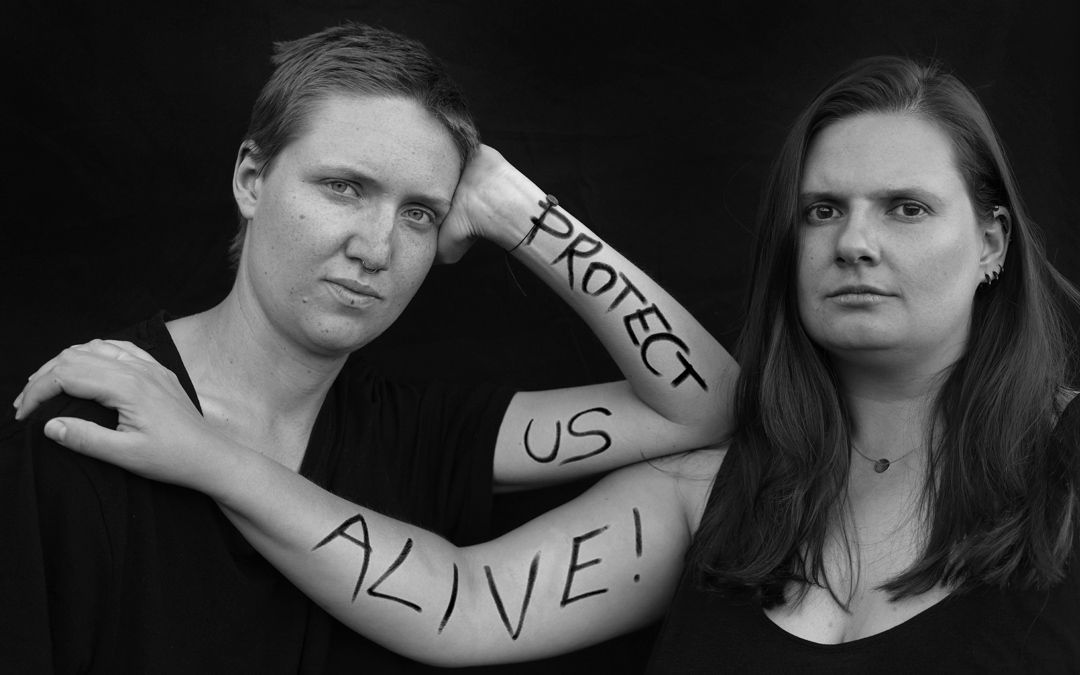 s/w-Bild: Zwei Frauen schauen selbstbewußt in die Kamera. Auf ihre Arme ist in schwarzen Großbuchstaben geschrieben: Protect us alive!