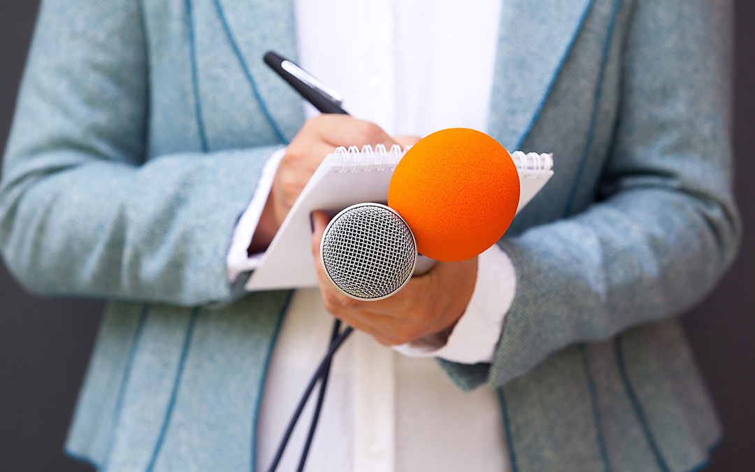 Frau hält zwei Mikrofone, eines ist orange, und Block auf dem sie schreibt