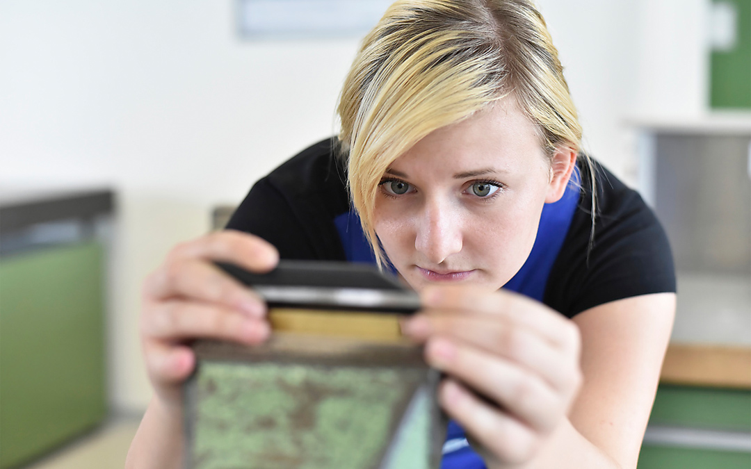 Junge blonde Frau schaut konzentriert an einer Werkbank auf ein Metallstück