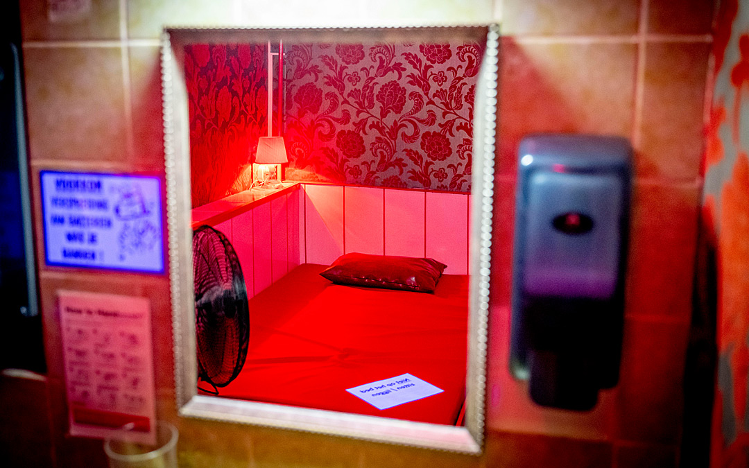 Blick durch ein Fenster auf ein leeres Bett in einem Bordell, rot angestrahlt.