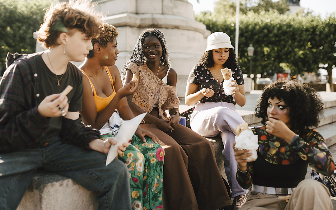 Fünf junge Frauen sitzen auf einem Mäuerchen unter einer Statue und plaudern beim Essen