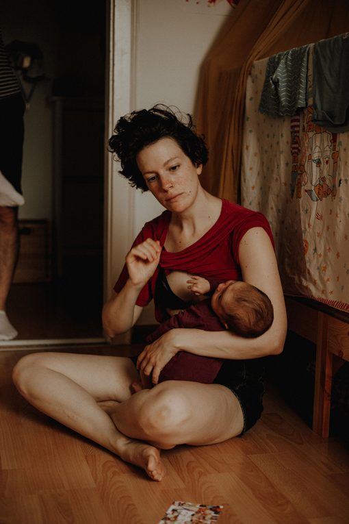 Kurz vorm Stillen: Müde Mutter sitzt mit Baby auf dem Fußboden, ihr Shirt ist halb hochgerutscht.