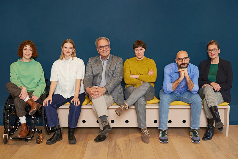 Gruppenbild vor einer blauen Wand. Frei Frauen und zwei Männer sitzen auf einer Bank, eine Frau links sitzt im Rollstuhl. Alle schauen lächelnd in die Kamera.