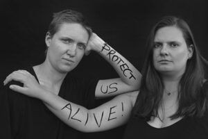 Zwei Frauen legen ihre Arme übereinander. Auf die Haut geschrieben steht: Protect us alive!