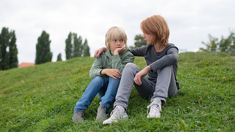 Zwei Jungen etwa 10 Jahre alt, sitzen auf einer Wiese, einer ist ein wenig traurig.